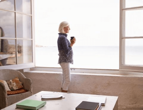 Woman standing in front of large, open window looking at the sea Woman standing in front of large, open window looking at the sea